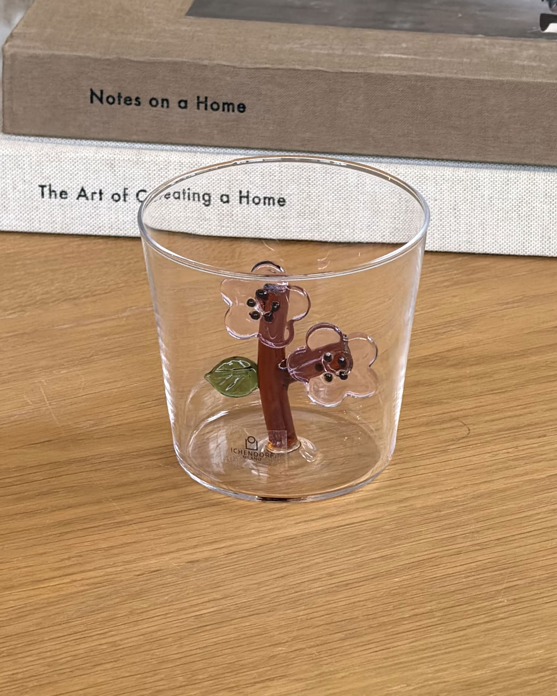 A glass of water in the Ichendorf Milano Botanica Tumbler - Peach Blossom sits on a wooden table with PROJECT JAPAN, a white clock, beige sofa, and colorful cushion, creating a serene scene inspired by Ichendorf Milano’s floral design.