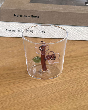 A glass of water in the Ichendorf Milano Botanica Tumbler - Peach Blossom sits on a wooden table with PROJECT JAPAN, a white clock, beige sofa, and colorful cushion, creating a serene scene inspired by Ichendorf Milano’s floral design.