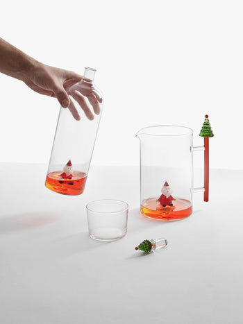 A hand pours orange liquid from the Ichendorf Milano Christmas Tales Bottle by Ichendorf Milano into a glass. On the white table are a clear pitcher with more orange liquid, the festive bottle inside, a tree-topped stirrer, and a glass.