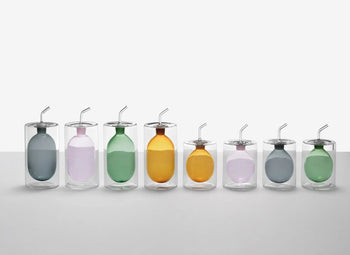 Eight transparent glasses with metal straws and colorful balloons are arranged in a row against a plain white background, echoing the modern, sleek design of the Ichendorf Milano Cilindro Oil Bottle by Ichendorf Milano.