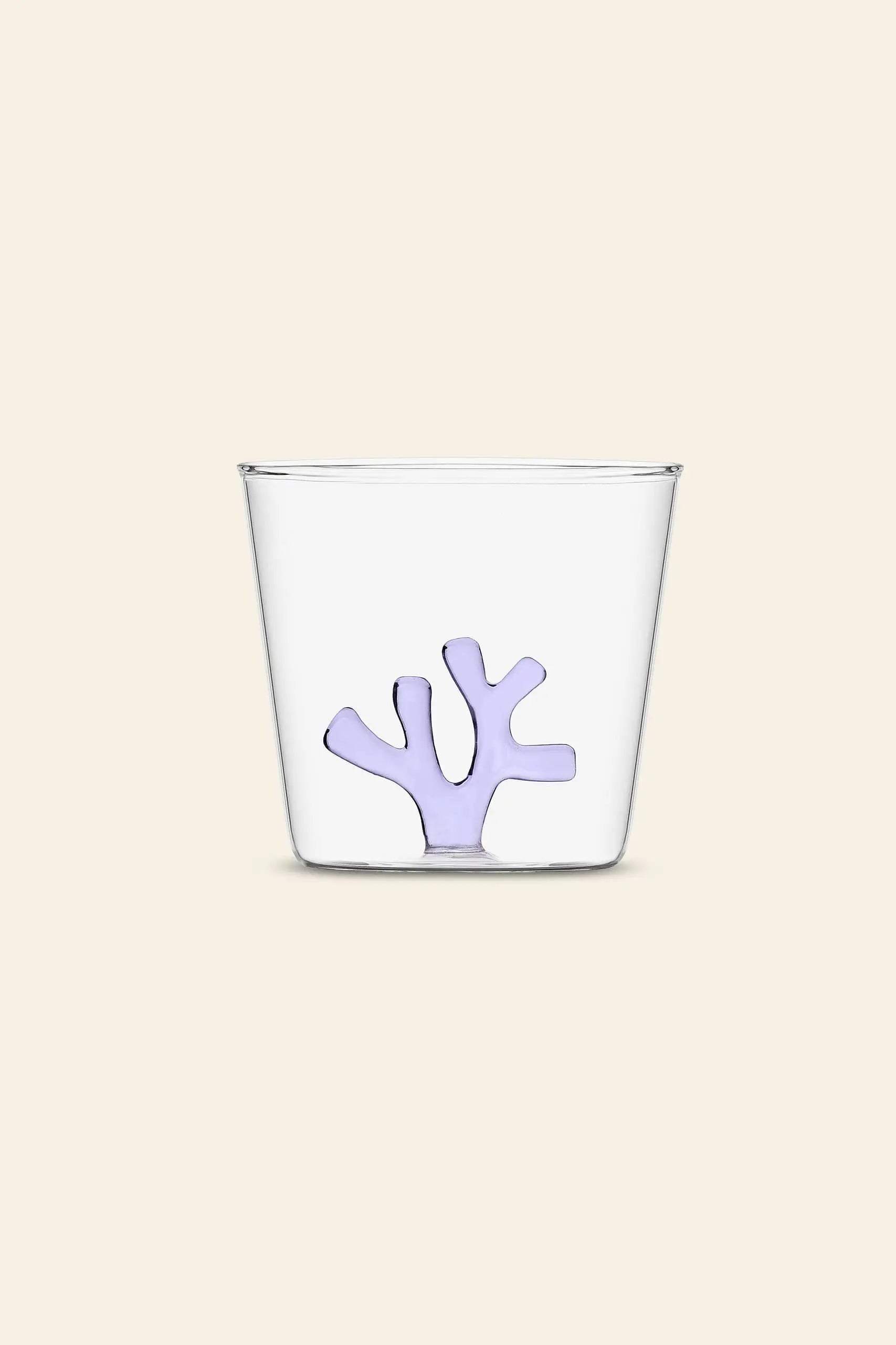 The Ichendorf Milano Coral Reef Tumbler by Ichendorf Milano is a clear glass with a small, abstract lavender coral-shaped design at the bottom, inspired by underwater ecosystems and set against a plain light beige background.
