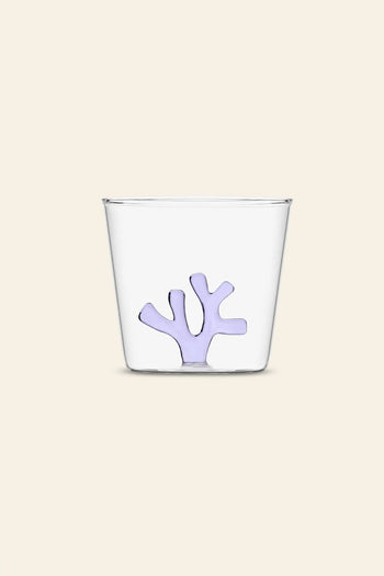 The Ichendorf Milano Coral Reef Tumbler by Ichendorf Milano is a clear glass with a small, abstract lavender coral-shaped design at the bottom, inspired by underwater ecosystems and set against a plain light beige background.