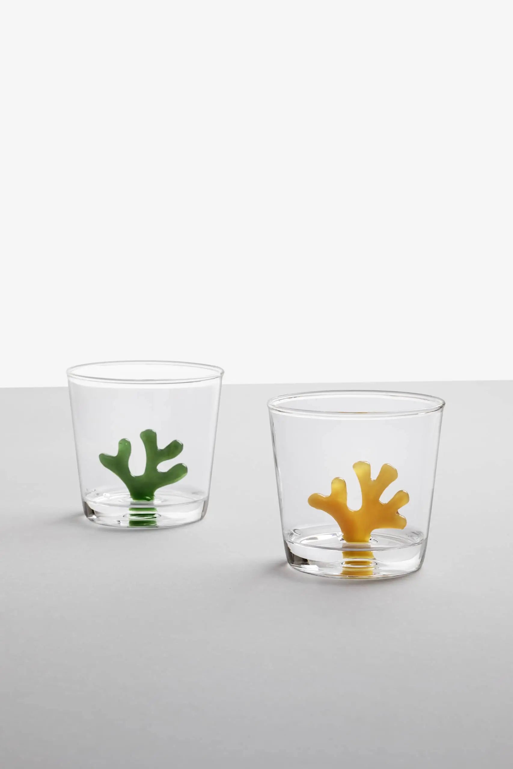 Two Ichendorf Milano Coral Reef Tumblers sit on a gray surface—a clear glass design by Ichendorf Milano, one with green coral and the other with yellow, each evoking vibrant underwater ecosystems against a plain white backdrop.