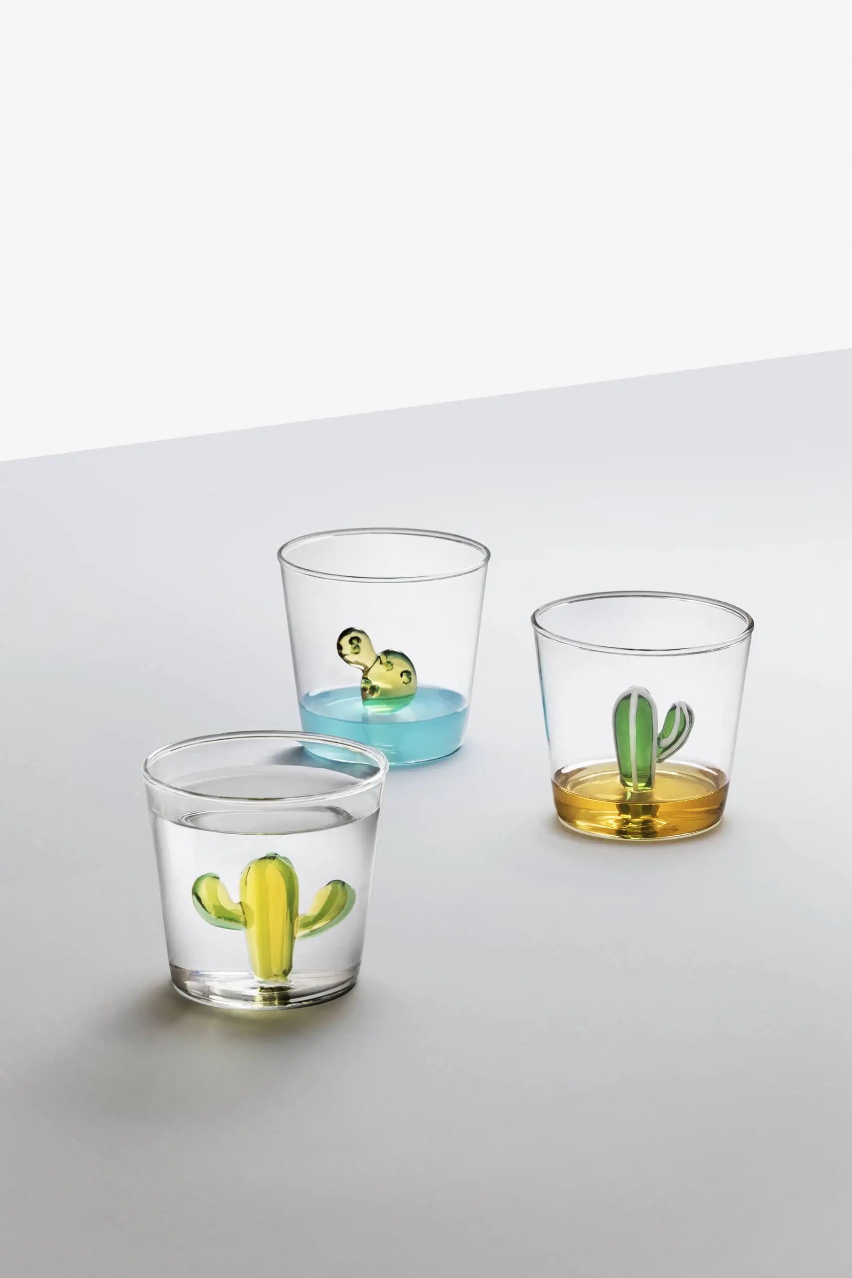 The Ichendorf Milano Desert Plants Tumbler set by Ichendorf Milano features three clear, handcrafted glasses with small cactus motifs—each displaying water, blue, or amber liquid—shown in a minimal scene against a white background.
