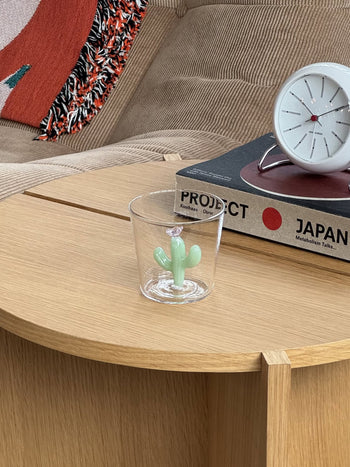 The Ichendorf Milano Desert Plants Tumbler by Ichendorf Milano, featuring a small green cactus inside clear handcrafted glass, sits on a light wooden table beside stacked books and a white clock with a beige couch in the background.