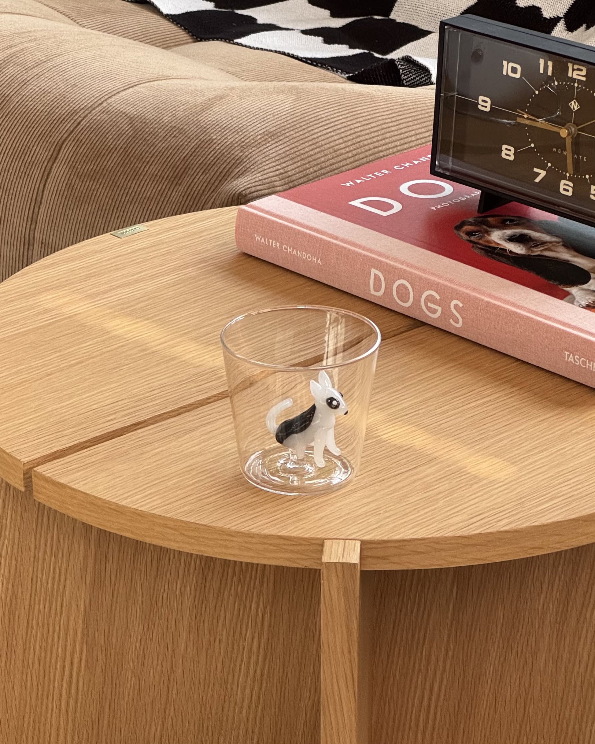 The Ichendorf Milano Dogs Tumbler - Bull Terrier, featuring a black and white dog figurine, rests on a round wooden table beside a book called DOGS and a clock, all displayed on a cozy, textured surface in warm tones.