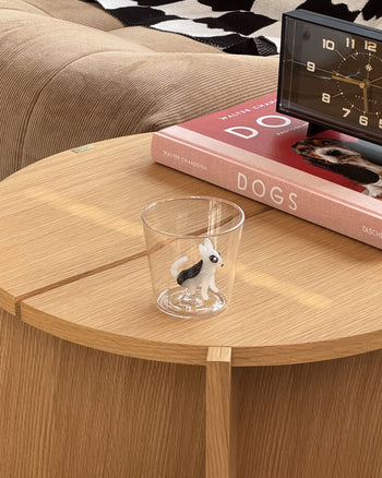 The Ichendorf Milano Dogs Tumbler - Bull Terrier, featuring a black and white dog figurine, rests on a round wooden table beside a book called DOGS and a clock, all displayed on a cozy, textured surface in warm tones.