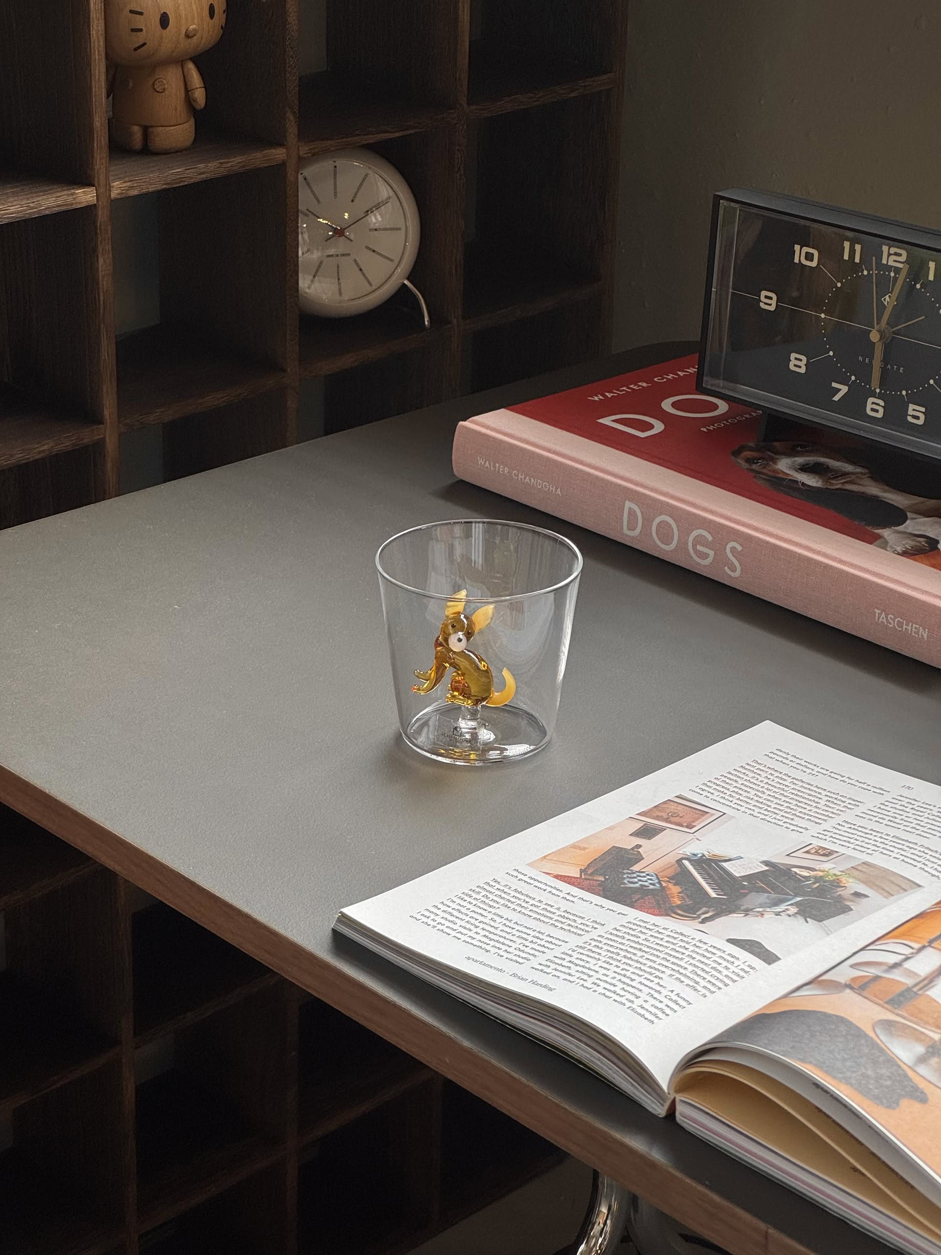 The Ichendorf Milano Dogs Tumbler - Chihuahua by Ichendorf Milano sits on a desk beside an open magazine, a book titled DOGS, a clock, and shelves with small items in the background.