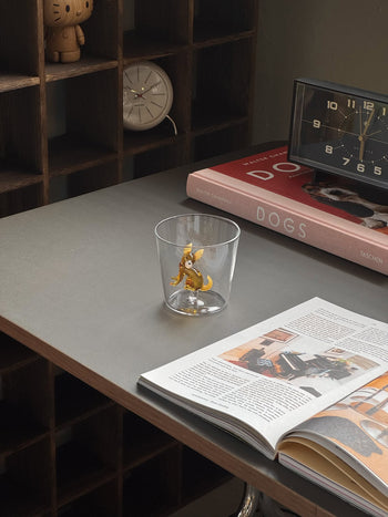 The Ichendorf Milano Dogs Tumbler - Chihuahua by Ichendorf Milano sits on a desk beside an open magazine, a book titled DOGS, a clock, and shelves with small items in the background.
