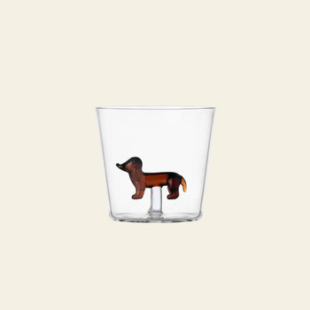 The Ichendorf Milano Dogs Tumbler - Dachshund by Ichendorf Milano features a clear glass design with a small brown dachshund figurine affixed to the inside bottom of the cup, set against a plain light background.