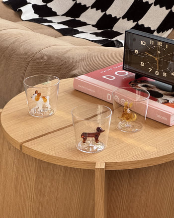 Three Ichendorf Milano Dogs Tumblers—each featuring a French Bulldog figurine—sit on a round table beside a black clock and two red books, with a beige sofa and black-and-white blanket in the background.