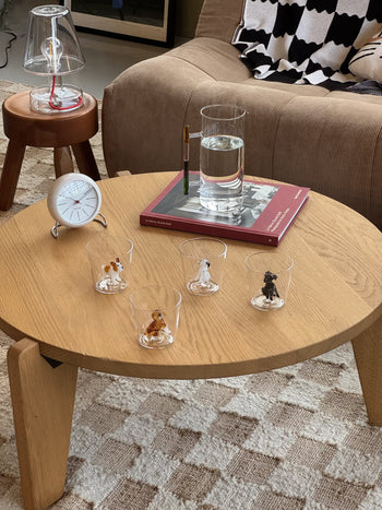 A round wooden coffee table showcases four Ichendorf Milano Dogs Tumbler - French Bulldog glass tumblers, a glass of water, a pen in a holder on a red book, and a white clock placed on an adjacent stool.