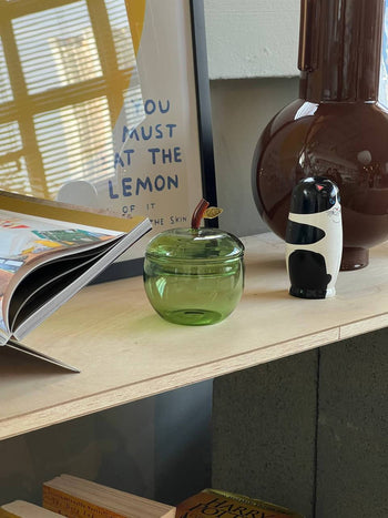 An Ichendorf Milano Fruits and Flowers Sugar Bowl rests on a wooden shelf beside an open book, a brown vase, assorted fruits, and framed art with the phrase YOU MUST AT THE LEMON.