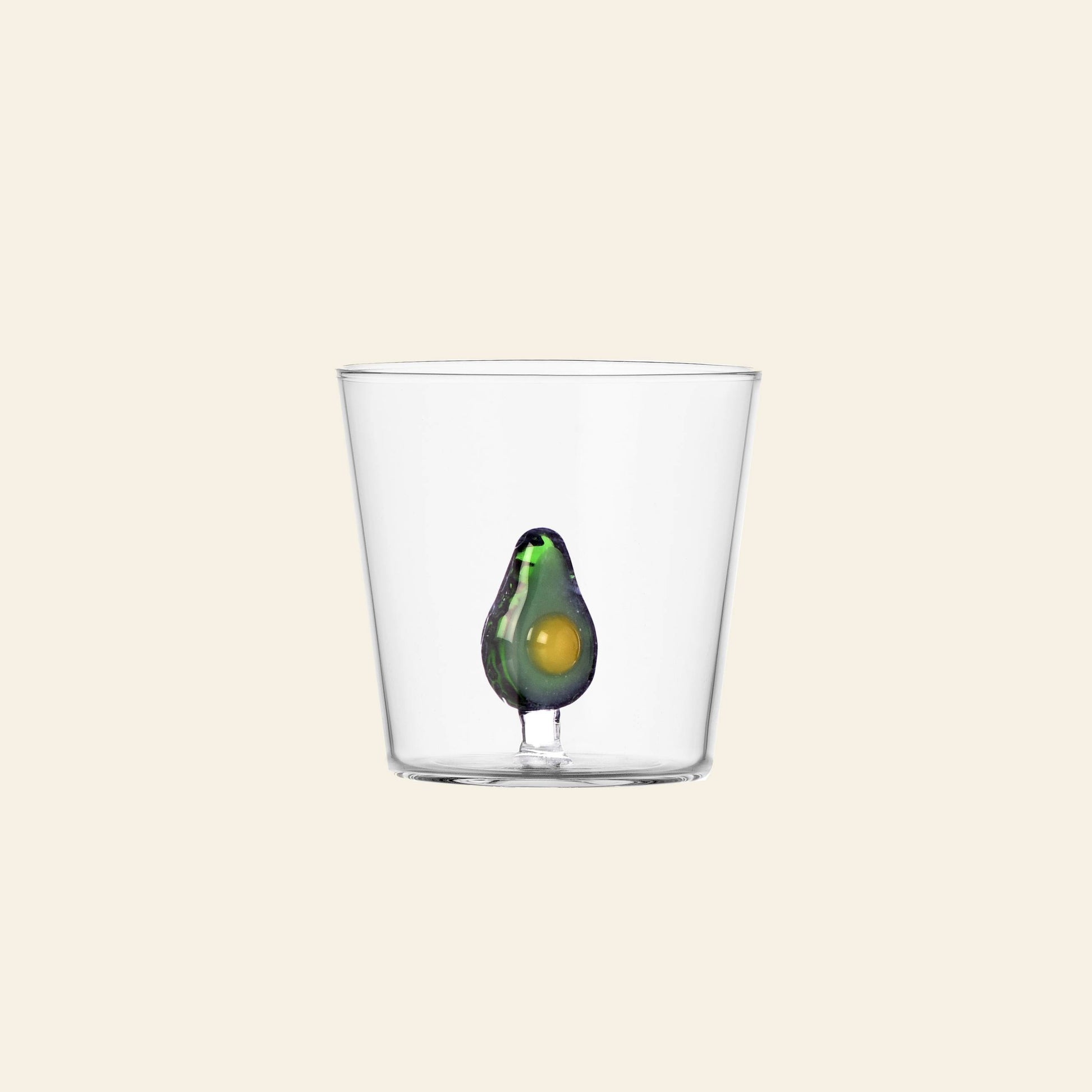 A clear Ichendorf Milano Fruits and Flowers Tumbler, featuring a small decorative avocado motif by Alessandra Baldereschi, displayed against a plain light beige background.