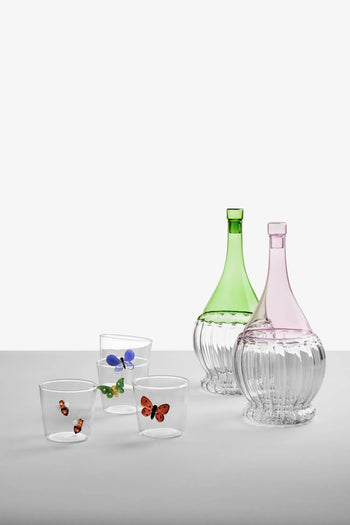 Two ornate glass decanters, one green and one pink, sit beside four Ichendorf Milano Garden Picnic Tumblers by Ichendorf Milano, each clear glass adorned with colorful butterfly and ladybug motifs, creating a whimsical garden vibe on a plain gray surface.