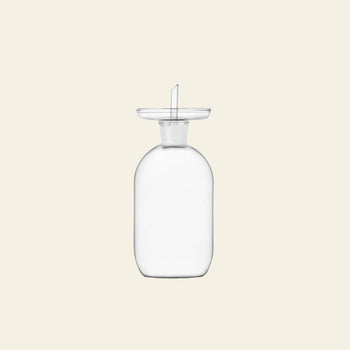 The Ichendorf Milano Lotus Oil Cruet Smooth Medium, a clear glass vessel with a wide neck and flat round stopper, stands upright against a light beige background—ideal for elevating any kitchen space.