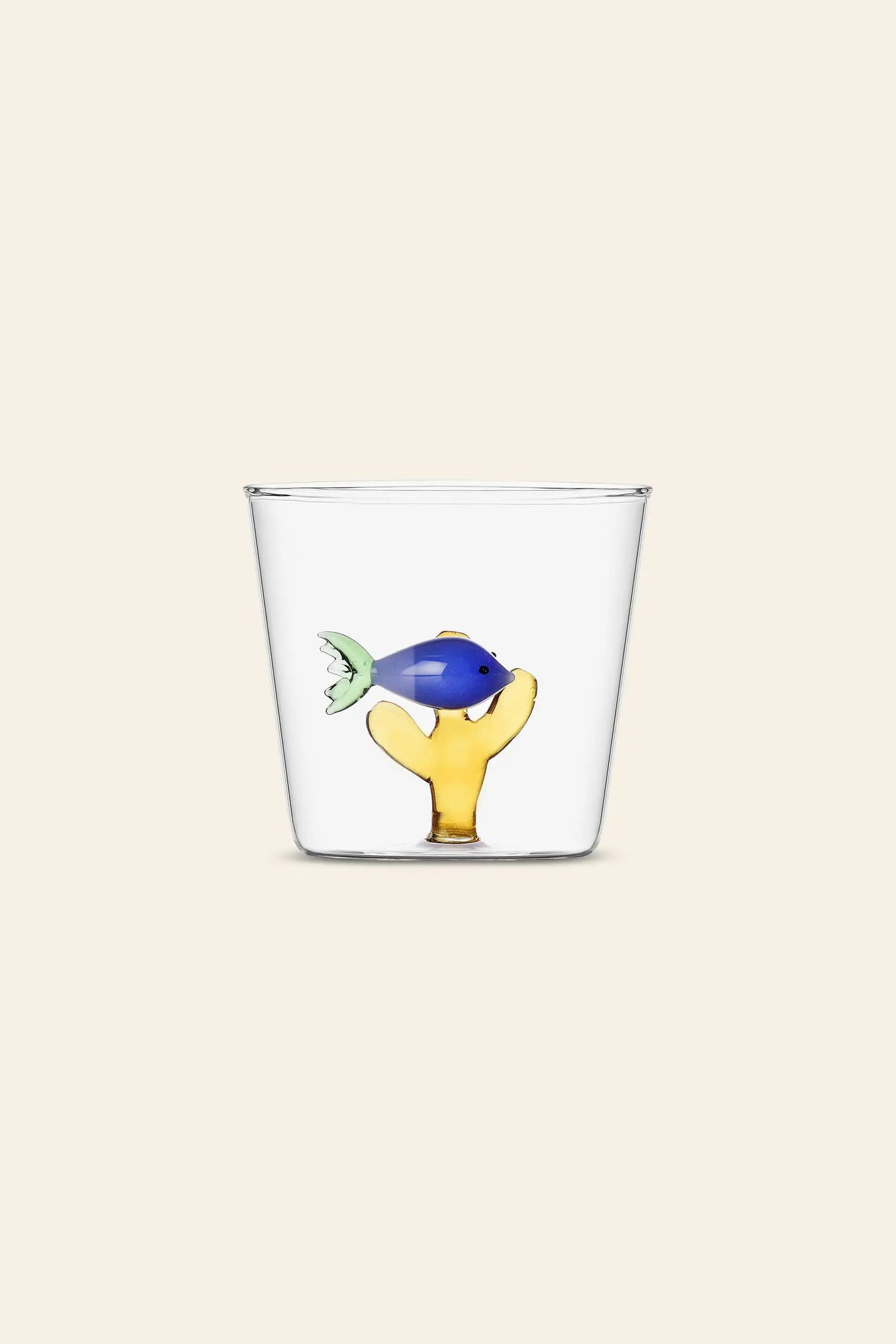 The Ichendorf Milano Marine Garden Tumbler by Ichendorf Milano is a clear glass cup featuring a small blue fish on a yellow coral-like base inside, set against a plain light beige background that highlights its aquatic charm.
