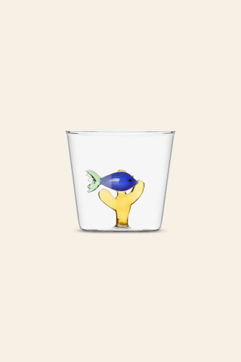 The Ichendorf Milano Marine Garden Tumbler by Ichendorf Milano is a clear glass cup featuring a small blue fish on a yellow coral-like base inside, set against a plain light beige background that highlights its aquatic charm.