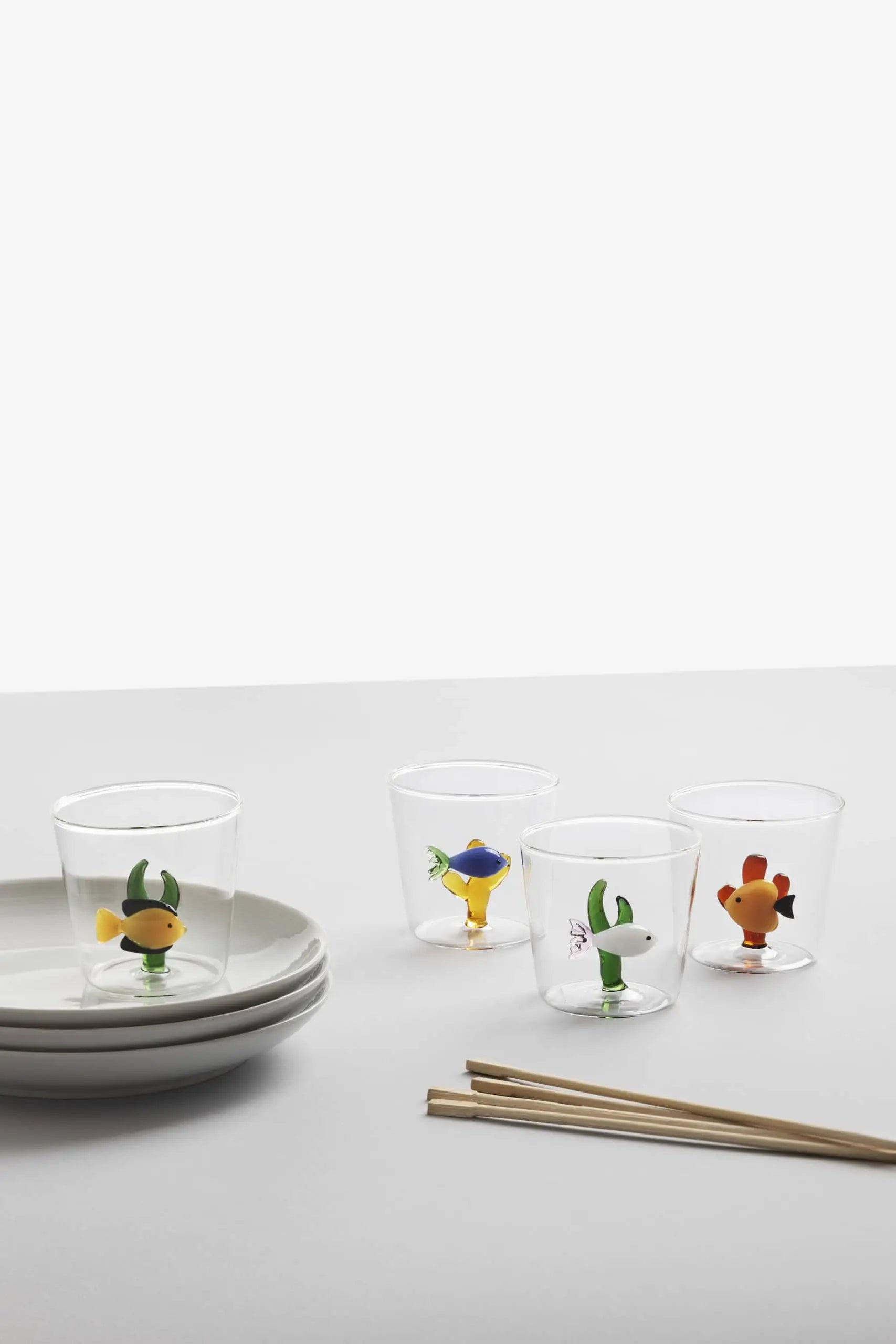 The Ichendorf Milano Marine Garden Tumblers—four clear glasses with colorful animal-shaped bases—by Ichendorf Milano are shown beside stacked white plates and chopsticks on a white surface with a plain background.