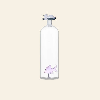 The Ichendorf Milano Tabby Cat Bottle by Ichendorf Milano is a clear glass bottle with a small purple fish figurine inside and a cat-shaped stopper on top, shown against a plain light beige background.