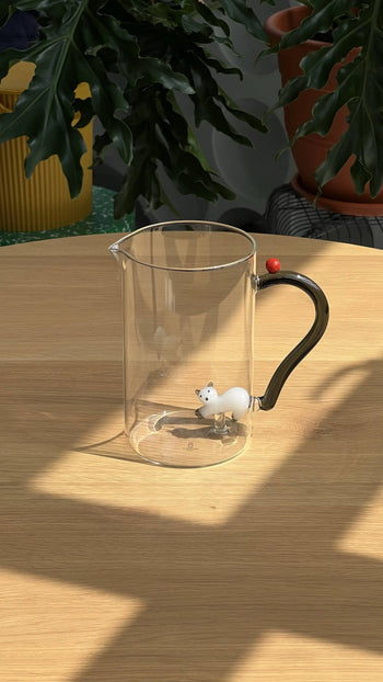 The Ichendorf Milano Tabby Cat Jug features a white cat with a smoke tail and berry inside clear glass. This handcrafted piece from Ichendorf Milano is displayed on a light wooden table, surrounded by green plants in dappled sunlight.