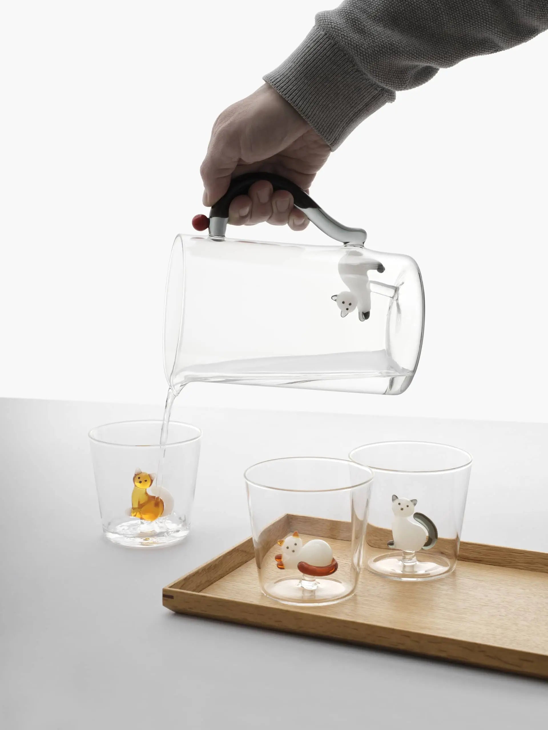 A hand pours water from the Ichendorf Milano Tabby Cat Jug—White Cat With Smoke Tail & Berry—into clear glasses with animal figurines, all on a light wooden tray; a perfect handcrafted gift for cat lovers by Ichendorf Milano.