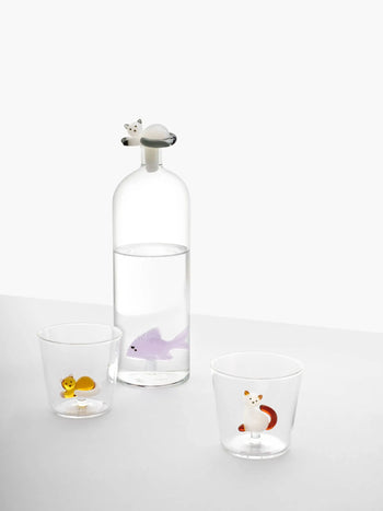 The Ichendorf Milano Tabby Cat Tumbler features hand-blown glass with animal figures—a yellow squirrel and a red fox—at the base, next to a water-filled bottle with a purple shark and a white cat-shaped stopper from Ichendorf Milano.