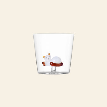 The Ichendorf Milano Tabby Cat Tumbler by Ichendorf Milano is a clear glass featuring a sculpted white cat with orange ears and tail at the bottom, set against a plain light beige background for a charming touch.