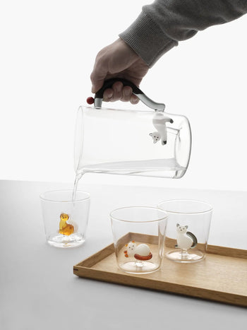 A hand pours water from an Ichendorf Milano pitcher into one of three Tabby Cat Tumblers by Ichendorf Milano, each featuring a duck, cat, or dog figurine at the bottom. The pitcher handle is adorned with a polar bear figurine.