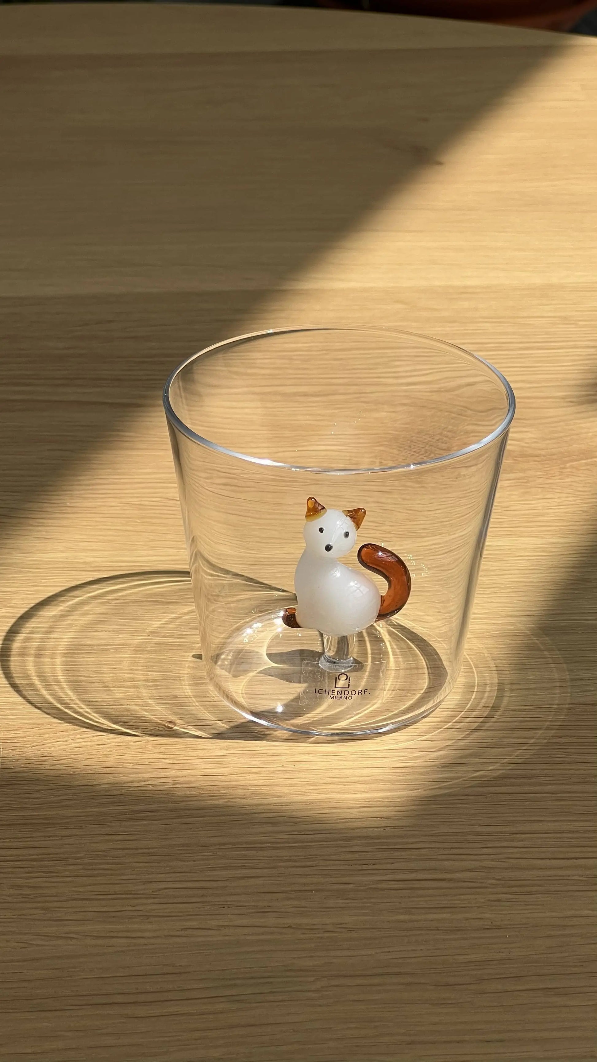 The Ichendorf Milano Tabby Cat Tumbler, a clear glass featuring a white and brown ceramic cat figurine, sits on a light wooden surface with sunlight casting shadows.