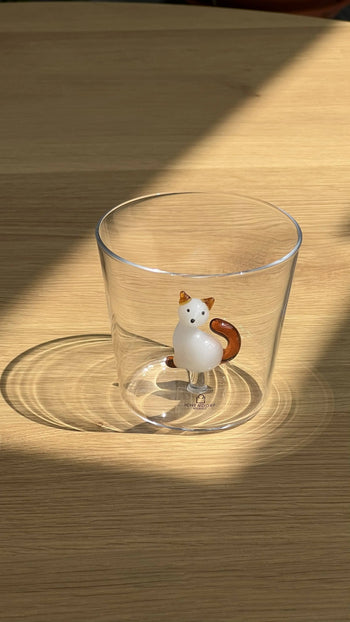 The Ichendorf Milano Tabby Cat Tumbler, a clear glass featuring a white and brown ceramic cat figurine, sits on a light wooden surface with sunlight casting shadows.