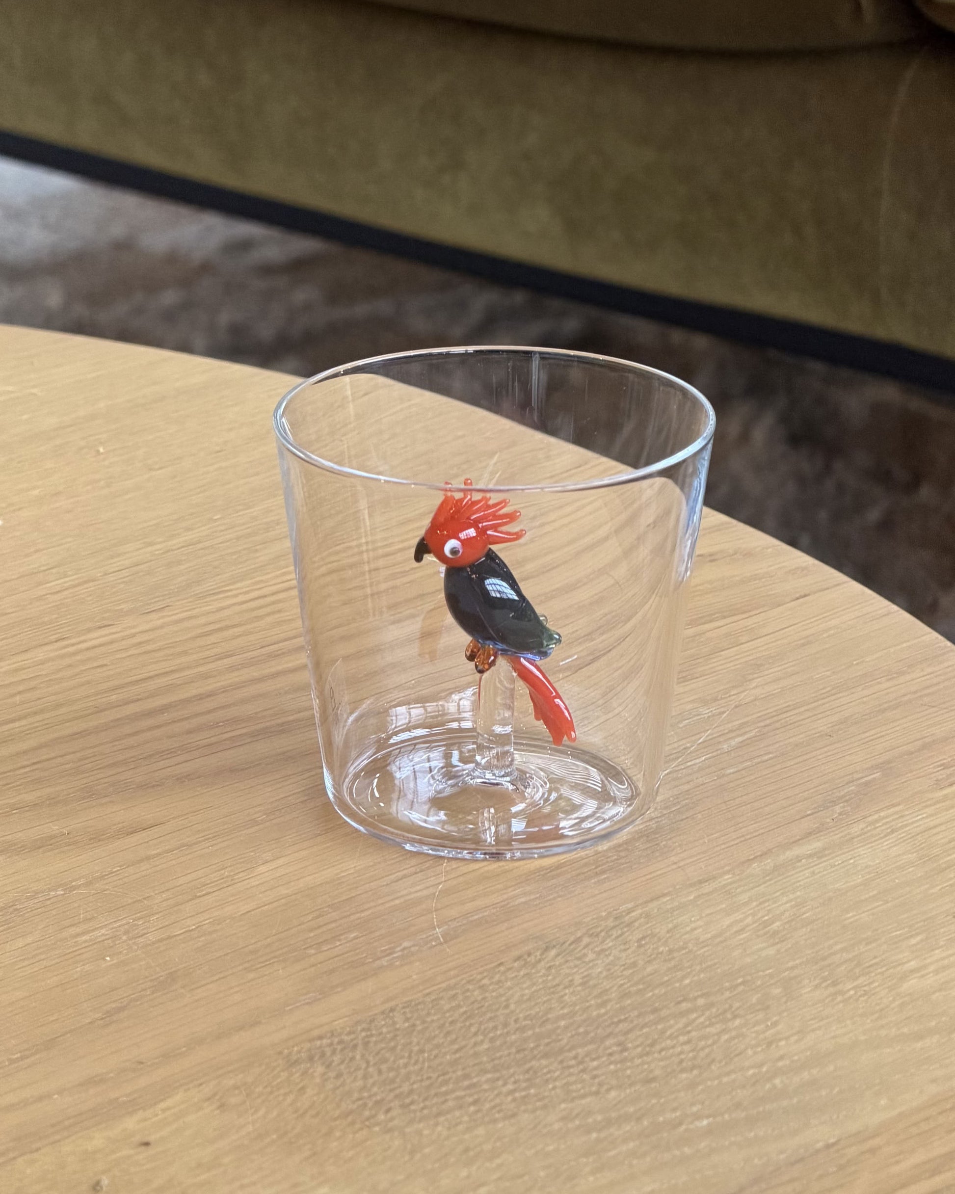 The Ichendorf Milano Talking Trees Tumbler - Red/Blue Cockatoo is a hand-crafted glass by Ichendorf Milano, featuring a decorative red and blue cockatoo figurine at its base, shown on a light wooden table with a green couch behind.
