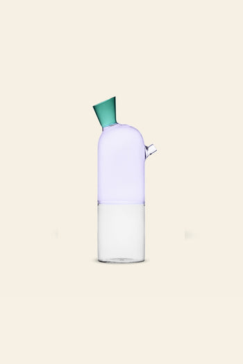 The Ichendorf Milano Travasi Bottle by Ichendorf Milano features a modern geometric design with a clear base, frosted light purple upper section, small side spout, and green angled cap, set against a plain beige background.