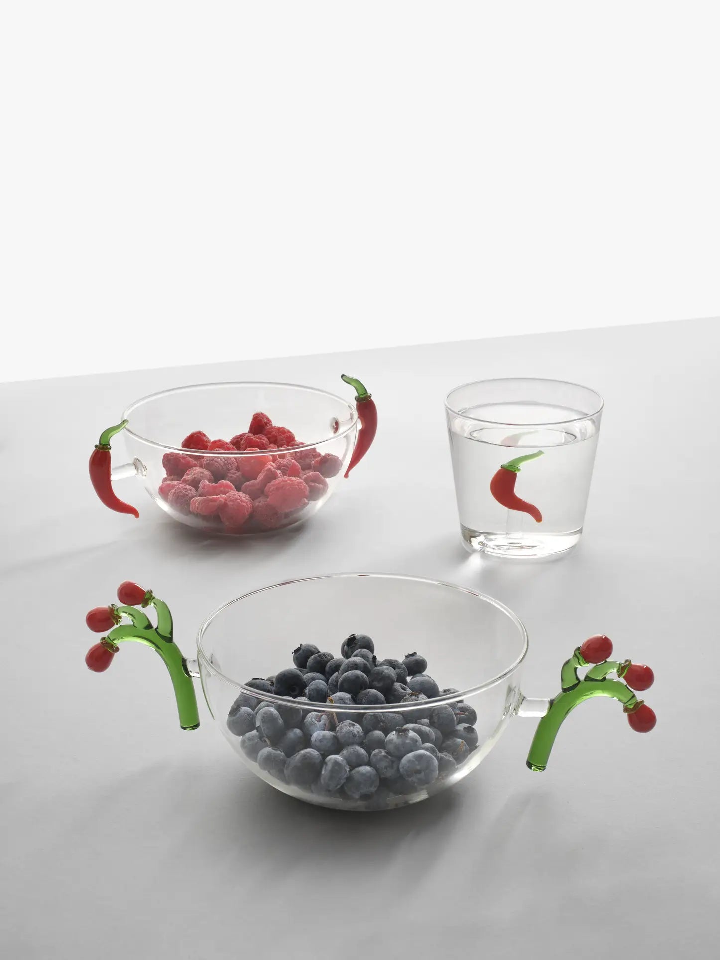 Three clear glass Ichendorf Milano Vegetables Bowls by Ichendorf Milano feature decorative handles shaped like chili peppers; they hold blueberries, raspberries, and water and display a painted red chili pepper motif.