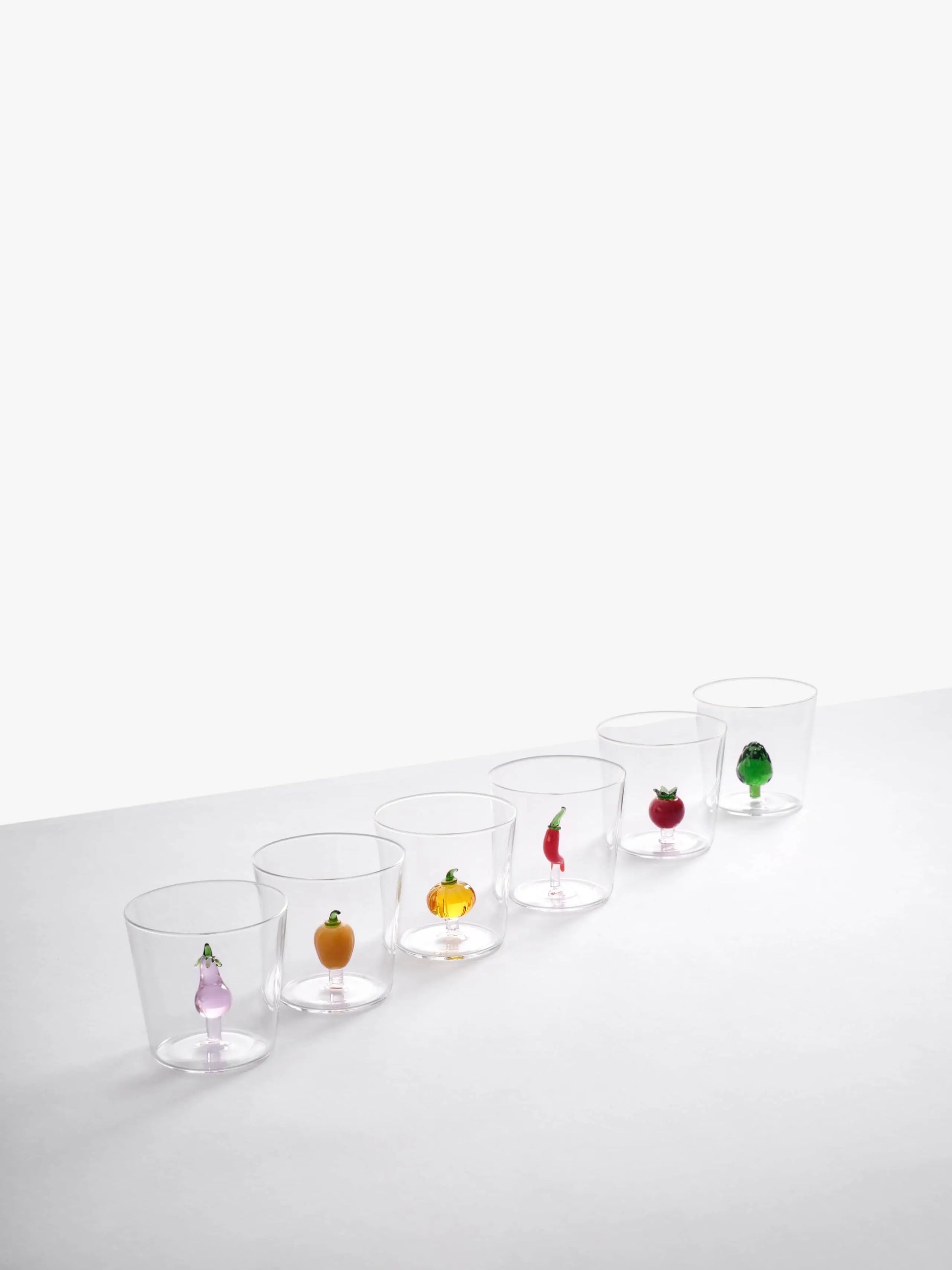 Six Ichendorf Milano Vegetables Tumblers are lined up, each showcasing a unique hand-blown glass figurine—onion, orange, pumpkin, chili pepper, eggplant, and green pepper—creating a charming display by Ichendorf Milano. White background.