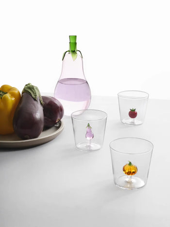A clear carafe with a green stopper sits beside a plate of yellow peppers and eggplants. Nearby are three Ichendorf Milano Vegetables Tumblers, each by Ichendorf Milano, featuring tiny glass vegetables inside.