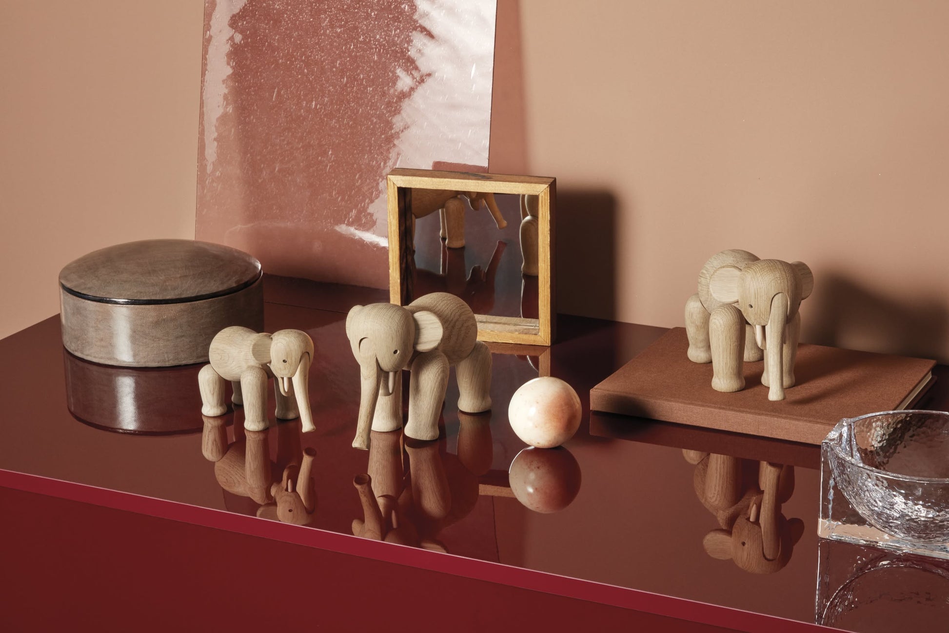 A glossy red surface displays three Kay Bojesen Elephant Mini figurines by Kay Bojesen, a round beige box, small framed artwork, tan ball, and textured glass bowl, set against a Danish-inspired beige and brown background.
