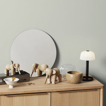 A wooden dresser with a round mirror, two Kay Bojesen Elephant Mini figurines by Kay Bojesen, a glass vase, a wooden bowl, a white table lamp with black base, a white dish with a yellow item, and keys on top.