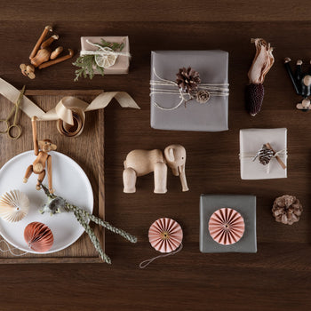 Flat lay of wrapped gifts with natural decorations, wooden toys like the Kay Bojesen Elephant Mini by Kay Bojesen, dried plants, scissors, ribbon, paper ornaments, and pinecones on a wooden table—celebrating Danish design.