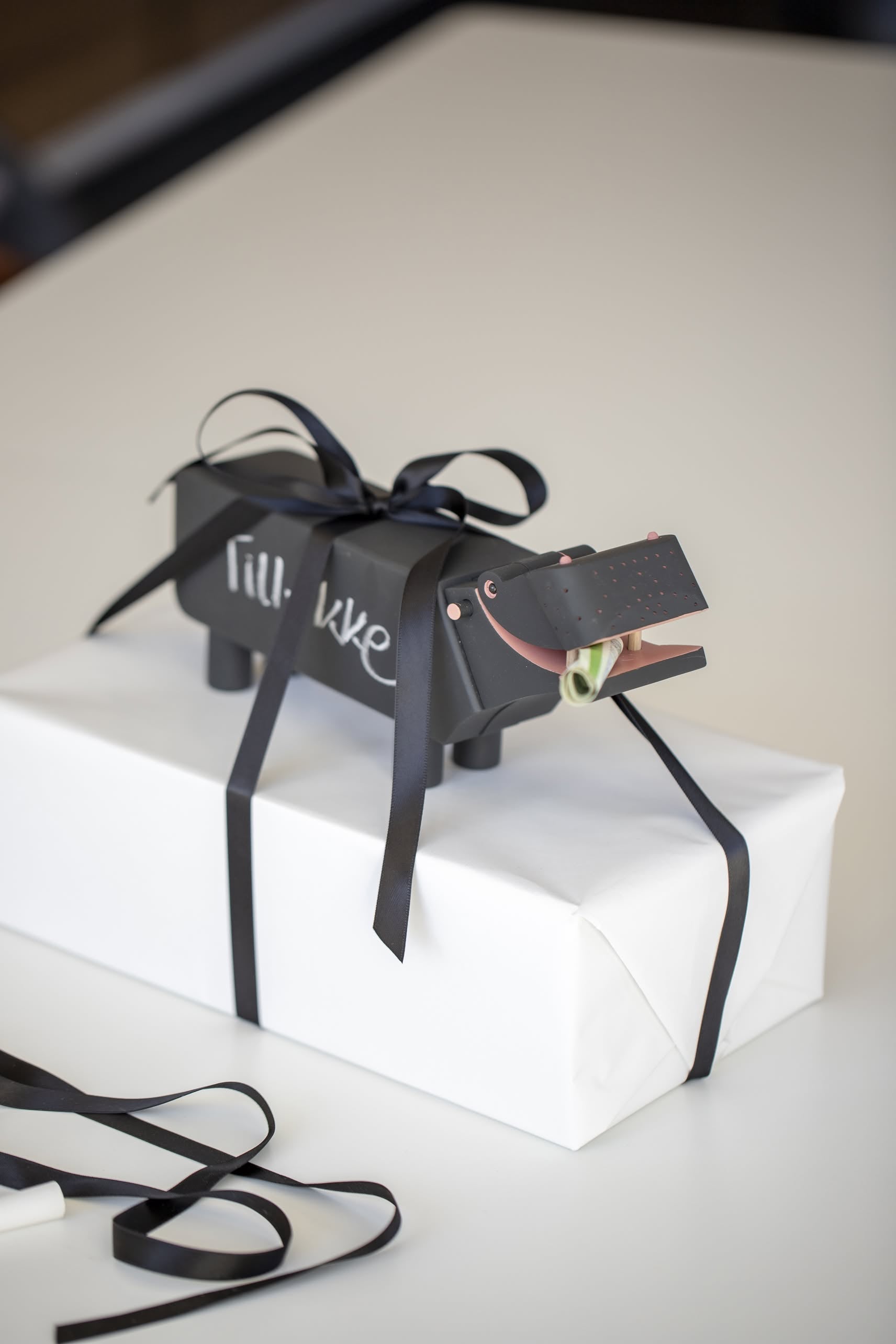 A white gift box wrapped with black ribbon, topped with a decorative Kay Bojesen Hippo figure featuring “lil’ luxe” on its side and a rolled bill in its mouth.