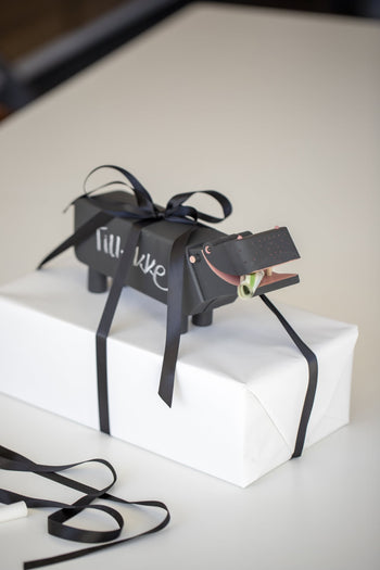 A white gift box wrapped with black ribbon, topped with a decorative Kay Bojesen Hippo figure featuring “lil’ luxe” on its side and a rolled bill in its mouth.