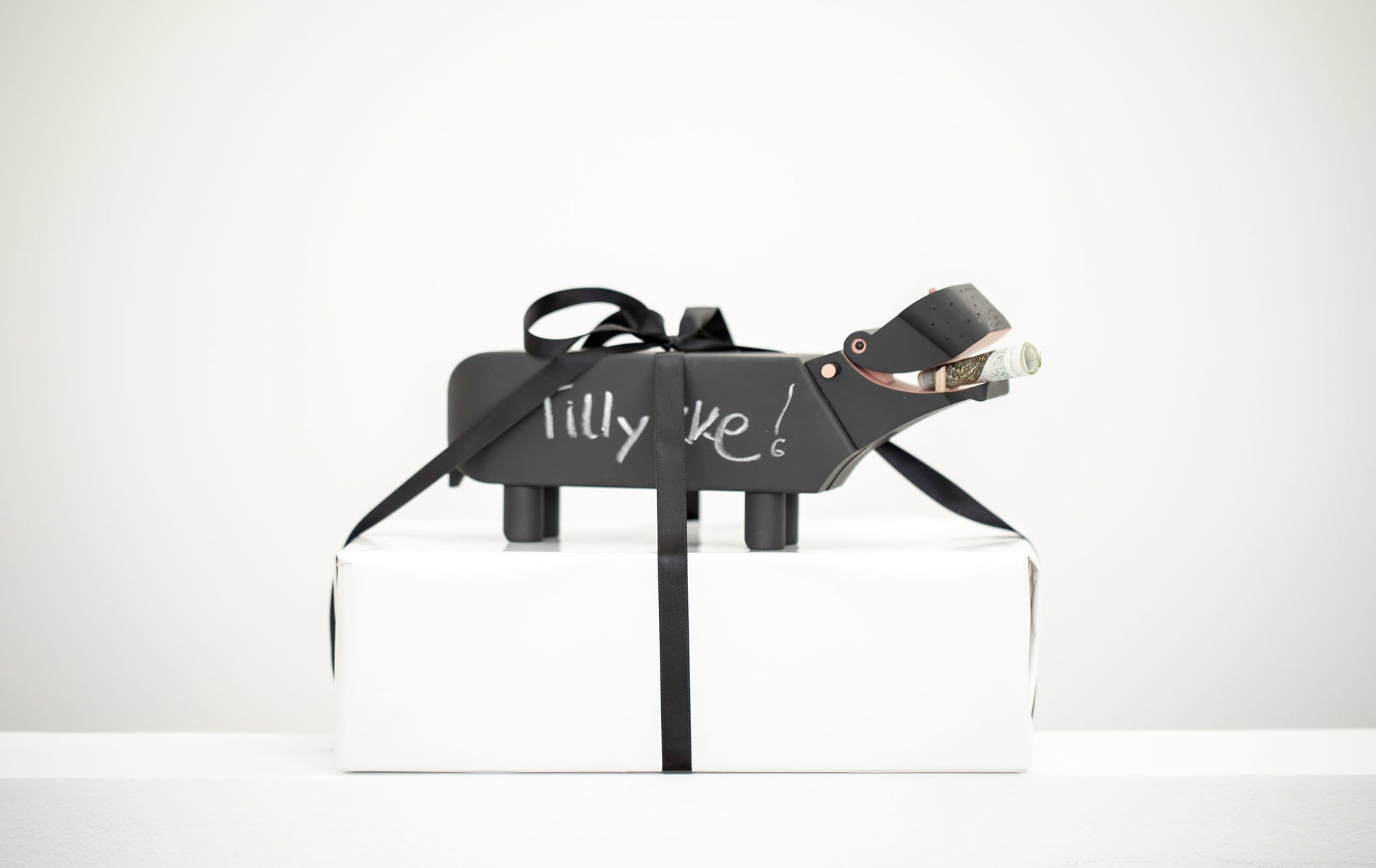 A small matte-black Kay Bojesen Hippo figurine by Kay Bojesen stands on a white box tied with a black ribbon, “Lilly Ike!” inscribed on its side, and holds a rolled-up bill in its mouth. Its finish recalls classic blackboard paint.