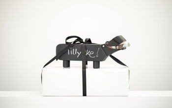 A small matte-black Kay Bojesen Hippo figurine by Kay Bojesen stands on a white box tied with a black ribbon, “Lilly Ike!” inscribed on its side, and holds a rolled-up bill in its mouth. Its finish recalls classic blackboard paint.
