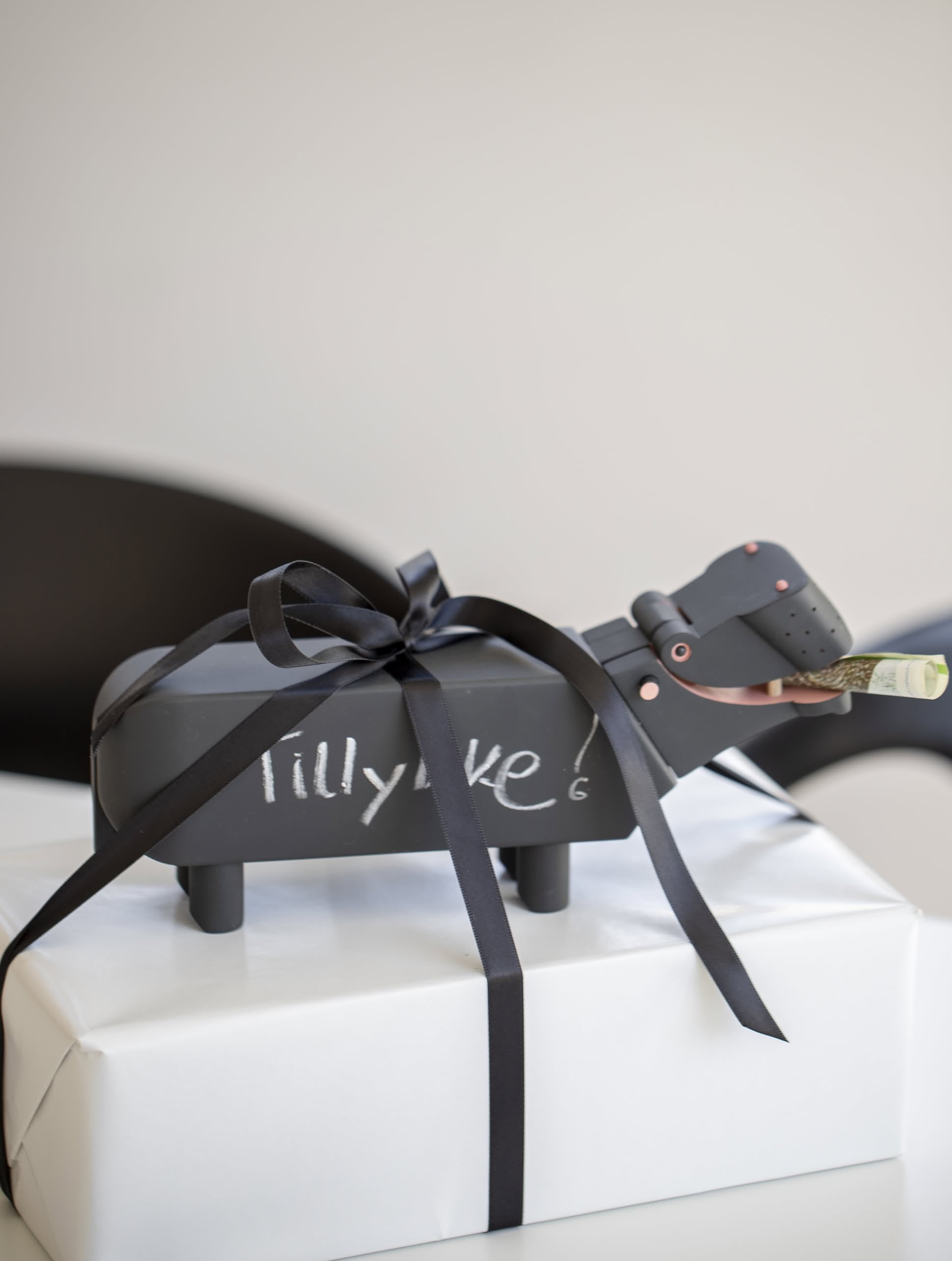A black Kay Bojesen Hippo gift box by Kay Bojesen, featuring “Tillykke!” and tied with a black ribbon, rests on a white present. A rolled-up bill sticks out of the hippos mouth, echoing the charm of playful wooden figures.