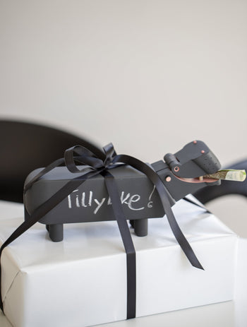 A black Kay Bojesen Hippo gift box by Kay Bojesen, featuring “Tillykke!” and tied with a black ribbon, rests on a white present. A rolled-up bill sticks out of the hippos mouth, echoing the charm of playful wooden figures.