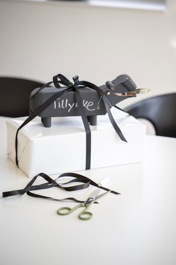 A white gift box with a black ribbon sits on the table, topped by the Kay Bojesen Hippo from Kay Bojesen—a wooden figure with blackboard paint and a note in its mouth. Nearby are green-handled scissors, extra ribbon, and two chairs.