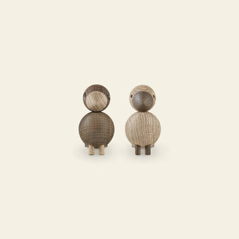 The Kay Bojesen Lovebirds Pair by Kay Bojesen features two wooden birds—one dark, one light—crafted with classic Danish design. Each has a simple rounded form and small legs, shown side by side on a plain, light background.