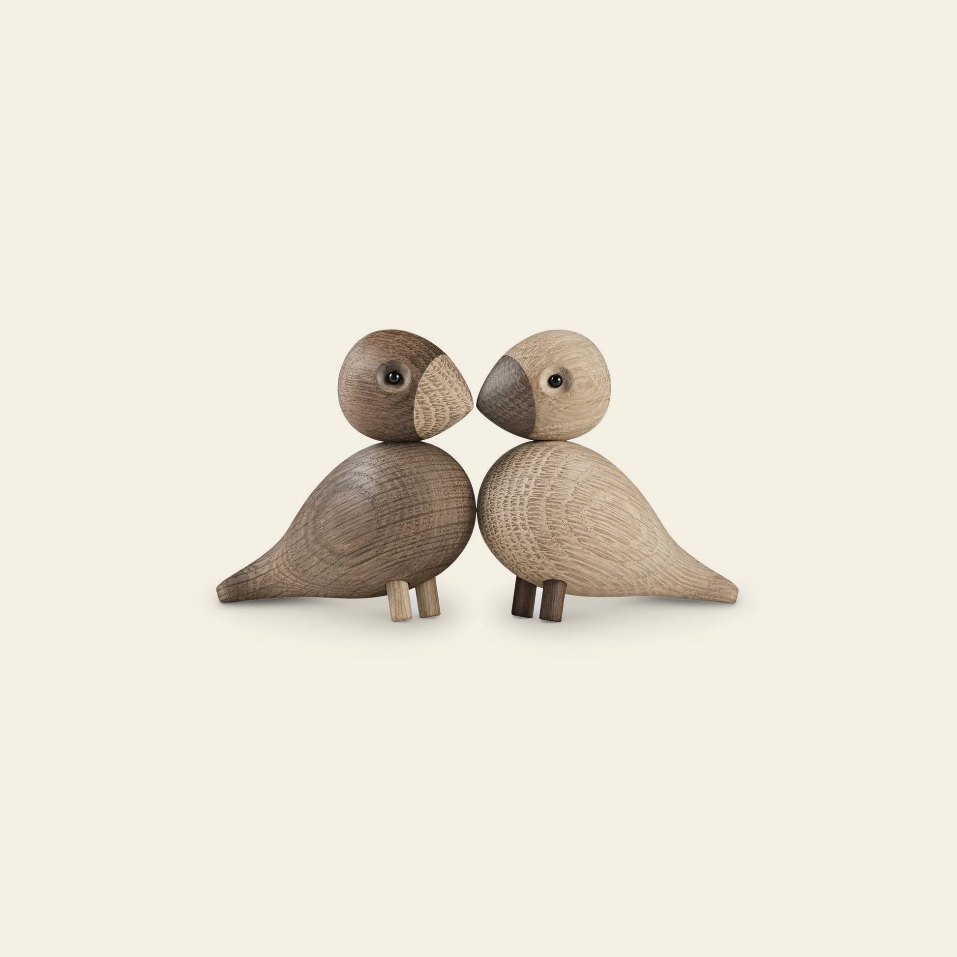 The Kay Bojesen Lovebirds Pair by Kay Bojesen features two wooden birds, one light and one dark, with simple rounded shapes and minimalistic details—a striking example of Danish craftsmanship against a light beige background.