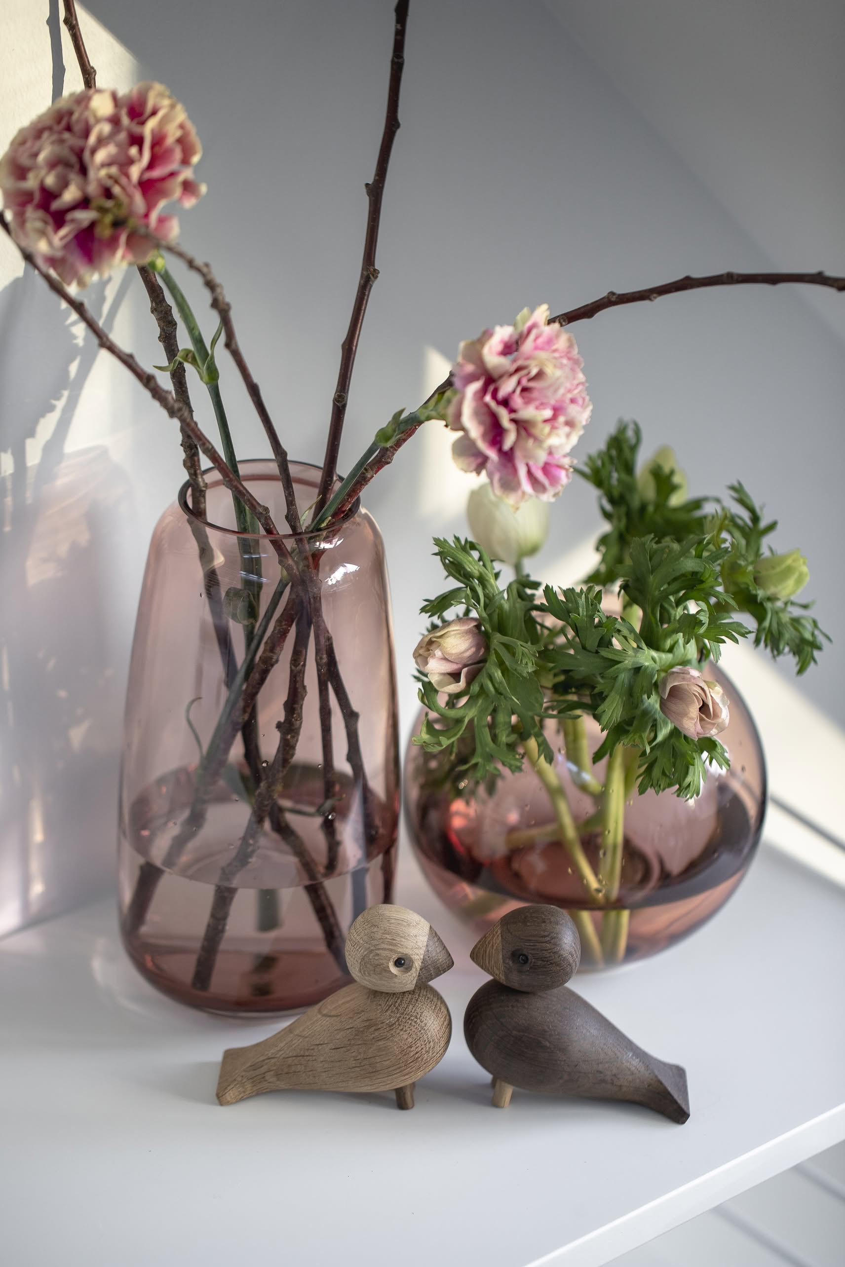 Two pink glass vases with flowers and branches rest on a white shelf. In front, the Kay Bojesen Lovebirds Pair by Kay Bojesen face each other, their Danish-inspired wooden charm adding warmth to the softly lit scene.