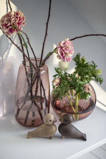 Two pink glass vases with flowers and branches rest on a white shelf. In front, the Kay Bojesen Lovebirds Pair by Kay Bojesen face each other, their Danish-inspired wooden charm adding warmth to the softly lit scene.